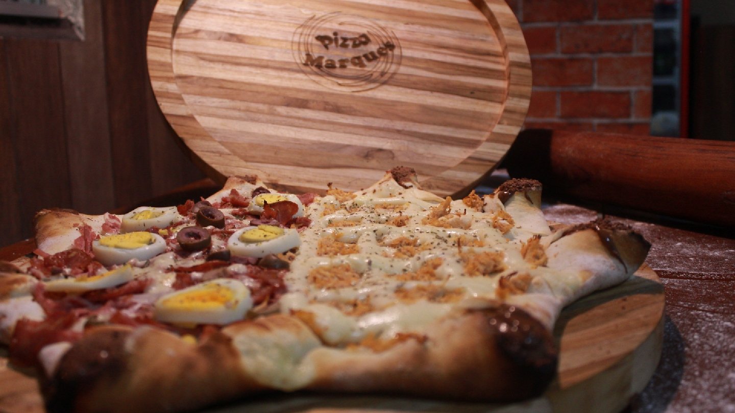 Pizza meio a meio na tábua de madeira com o logo Pizza Marques marcado a fogo ao fundo